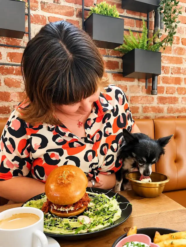 Guest with a pulled pork burger while her black chihuahua eats from a gold bowl.
